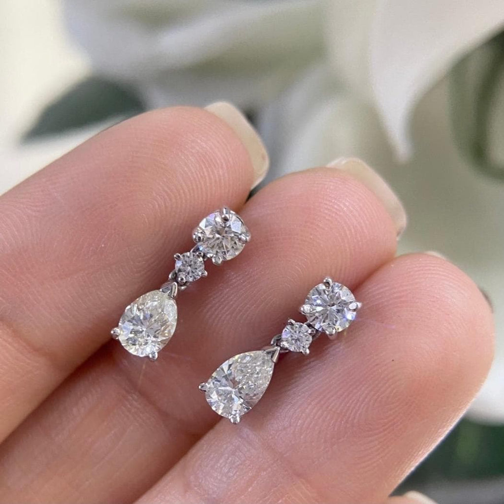 Woman holding white gold three stone diamond drop earrings. Top stone Round Cut diamond, middle stone smaller cut diamond, bottom stone pear shape diamond. (etsy image id: 6806049231)